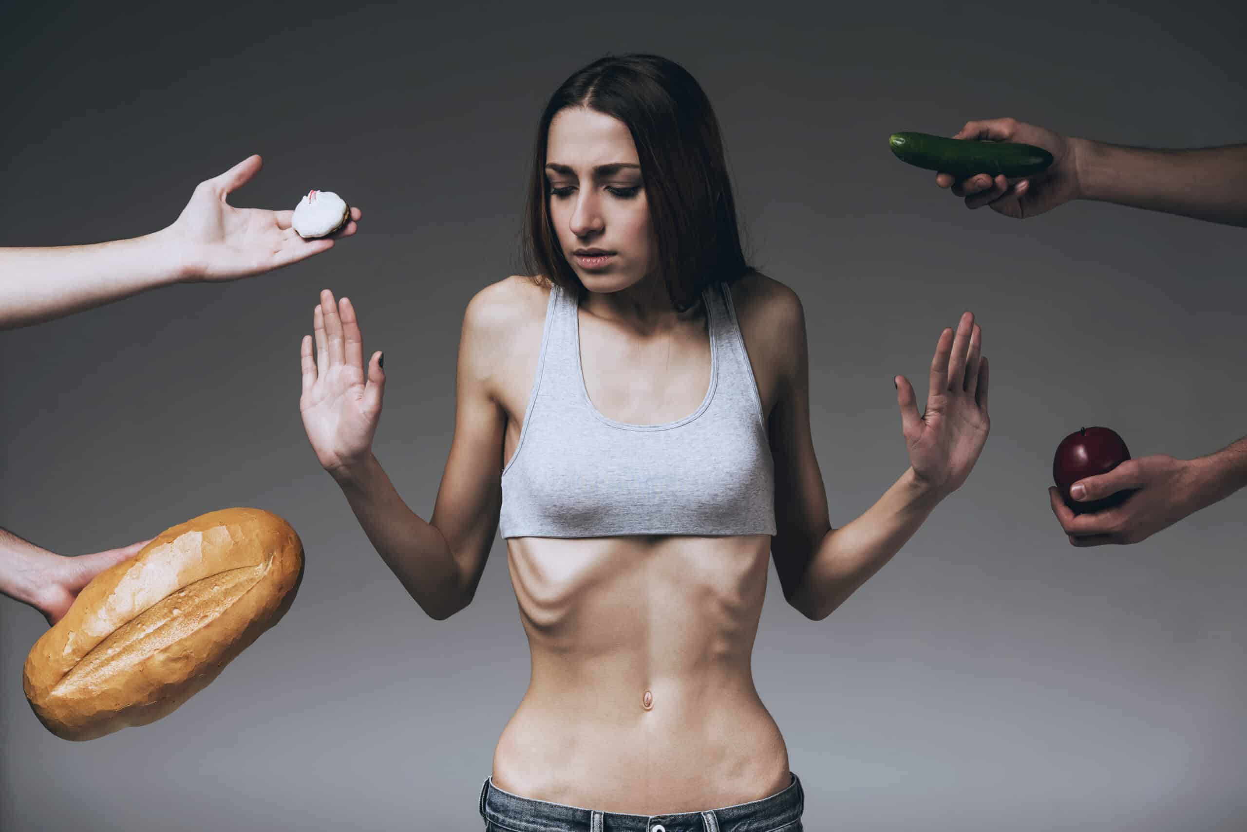 The image depicts a young woman, visibly emaciated, positioned centrally against a muted grey background. She is wearing a light grey tank top and dark denim jeans, emphasizing her extremely thin frame and prominent ribcage, with disembodied hands offering food — a stark depiction of struggle with eating and body image.
