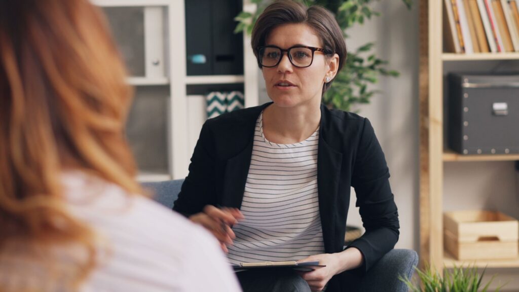 A therapist in glasses talks with a client in a room with bookshelves and plants