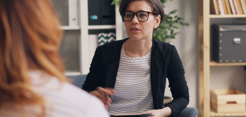 A therapist in glasses talks with a client in a room with bookshelves and plants